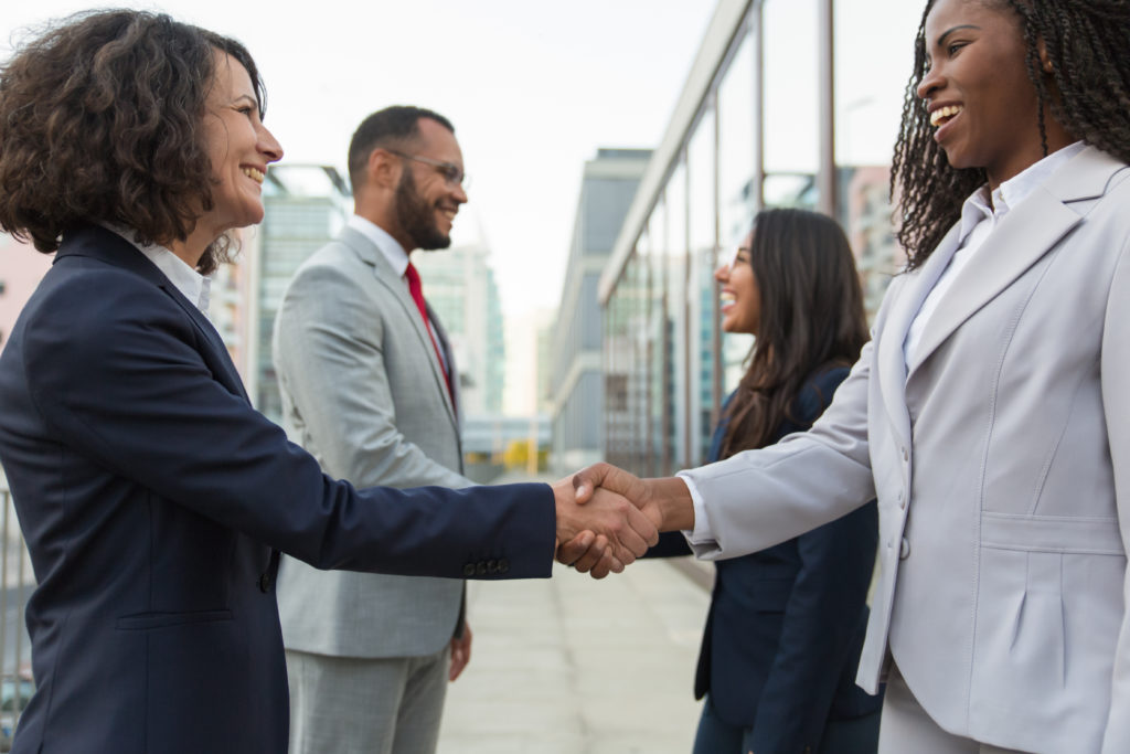 Happy confident diverse business partners meeting in city. Business man and women standing in urban street, shaking hands with each other. Conference concept