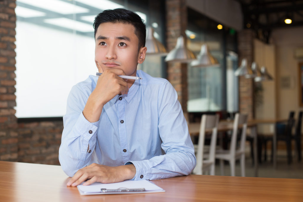 Closeup portrait of young Asian man thinking on project and sitting at table with clipboard and blurred cafe interior in background