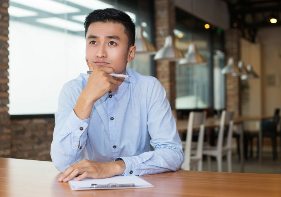 Closeup portrait of young Asian man thinking on project and sitting at table with clipboard and blurred cafe interior in background